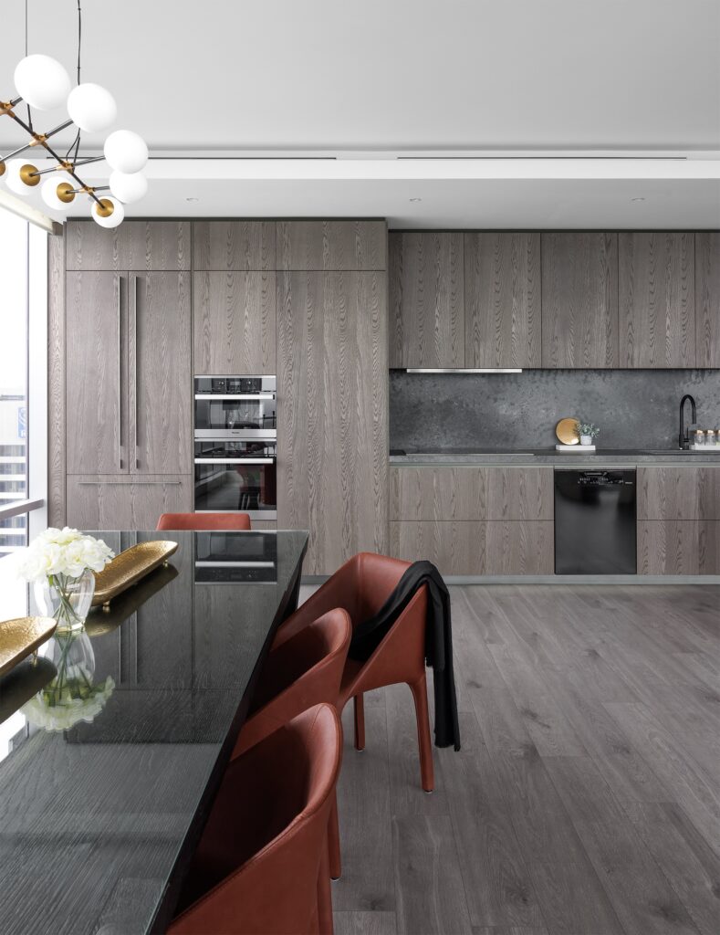 A modern kitchen with gray wood cabinets, a dark stone backsplash, built-in appliances, and a black countertop. Reddish-brown chairs line a dining table. A modern chandelier and a vase with white flowers decorate the space.