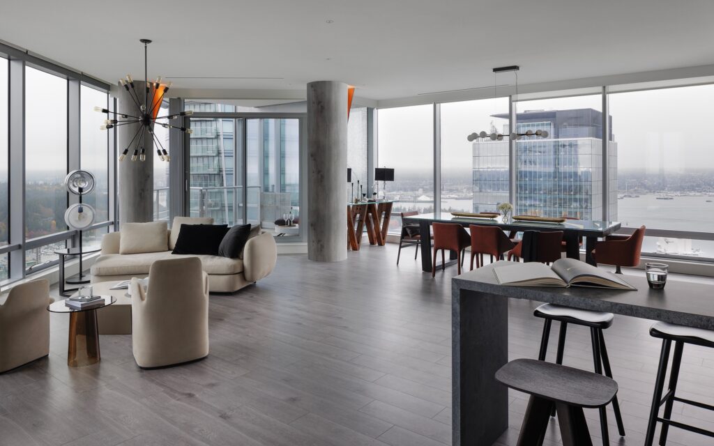 A modern, open-concept apartment with large floor-to-ceiling windows, neutral-toned furniture, a dining area with red chairs, a kitchen island with stools, and city and water views outside.