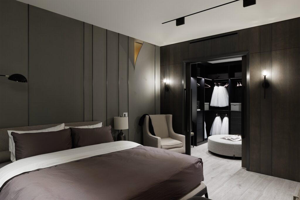 A modern bedroom with dark walls, a double bed with brown and white bedding, an armchair, and an open walk-in closet displaying several white dresses and a round ottoman. Soft lighting creates a cozy atmosphere.