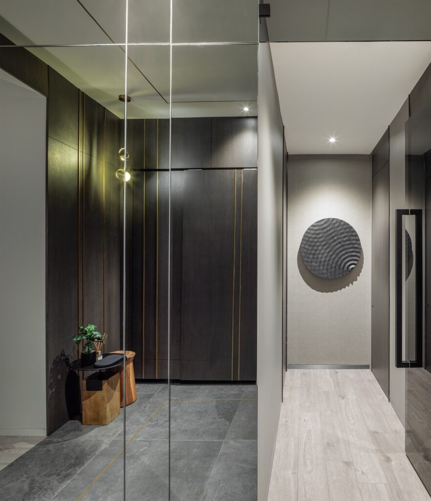 Modern hallway with dark wood paneling, a small wooden stool and plant, gray tile flooring, and a mirrored wall. A textured black circular wall art hangs on a light wall at the end of a light wood corridor.
