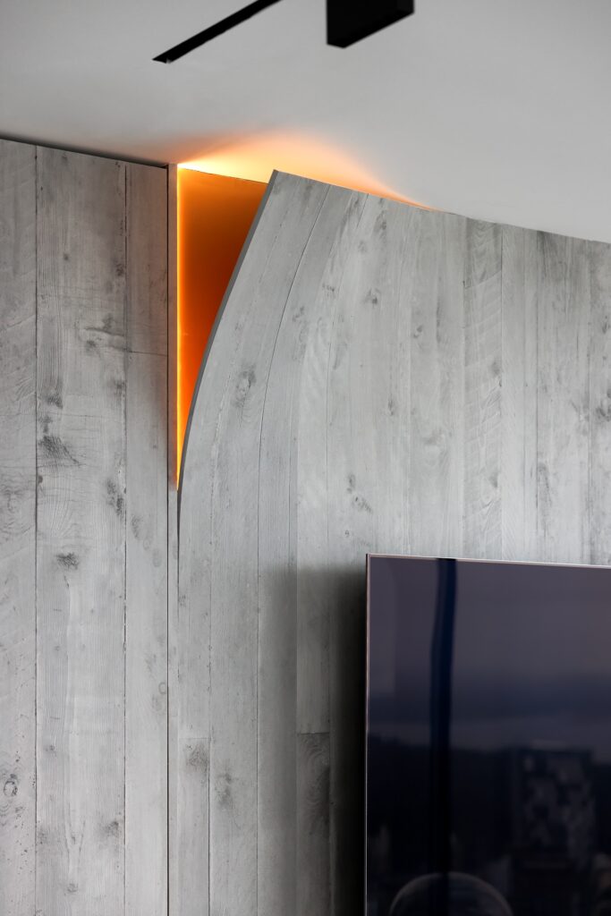 A close-up of a modern interior wall with light gray wood panels, featuring a curved section that reveals a strip of warm orange backlighting behind it and part of a flat-screen TV in the foreground.