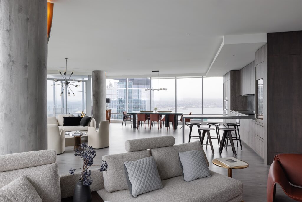 A modern, open-concept living space with large windows, light gray sofas, a dining table, dark wood kitchen cabinets, barstools, and city views in the background.