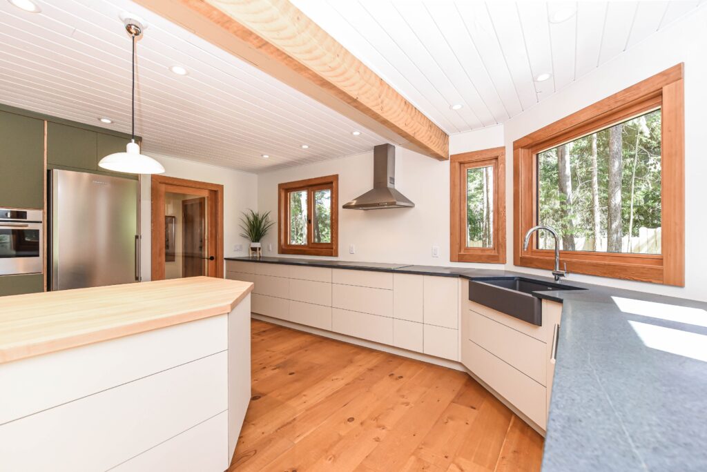 Modern kitchen with light wood floors, white cabinetry, dark countertops, stainless steel appliances, a farmhouse sink, and large windows with wooden frames letting in natural light.