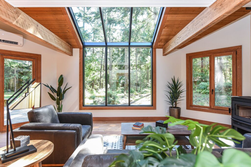 Modern living room with large glass windows and skylight, wooden beams, black leather sofas, green plants, and a view of a lush forest outside. Natural light fills the bright, cozy space.