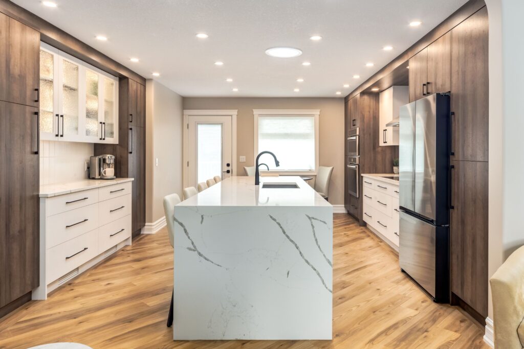 Modern kitchen with wood cabinets, white countertops, a marble island with a sink, stainless steel appliances, bar stools, and recessed lighting; the room has light walls and wood flooring.