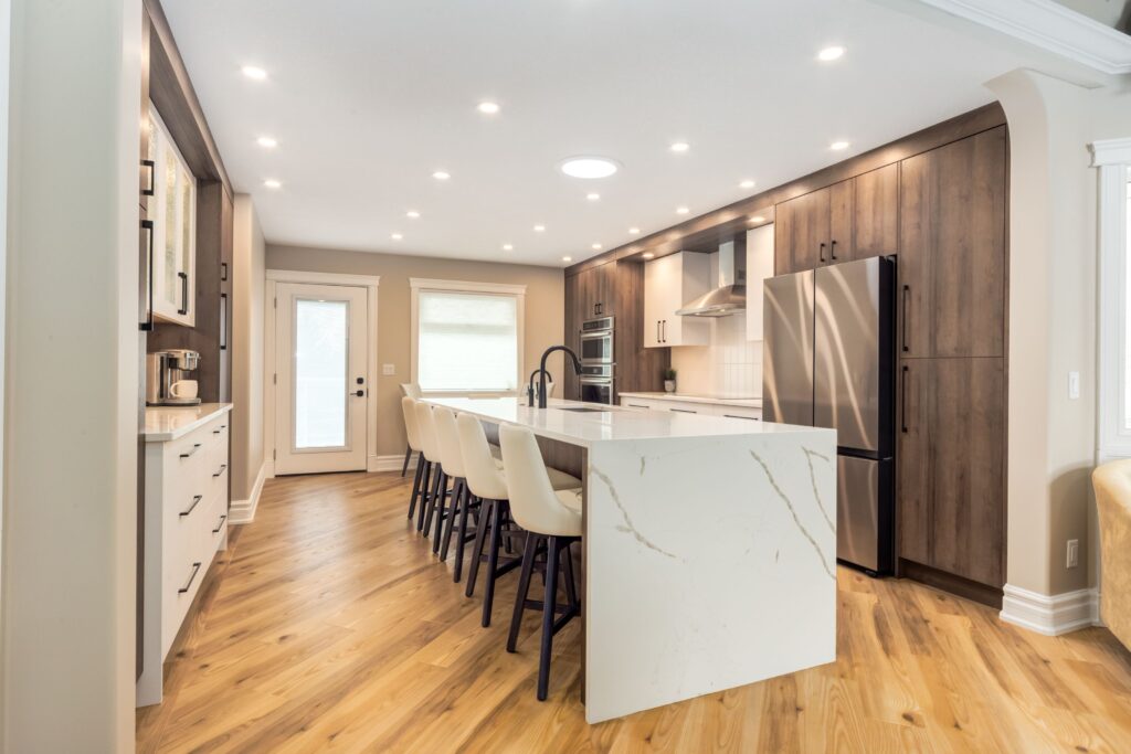 Modern kitchen with wood floors, white marble island, six white chairs, stainless steel appliances, and dark wood cabinets. Recessed ceiling lights brighten the space, and a glass door leads outside.