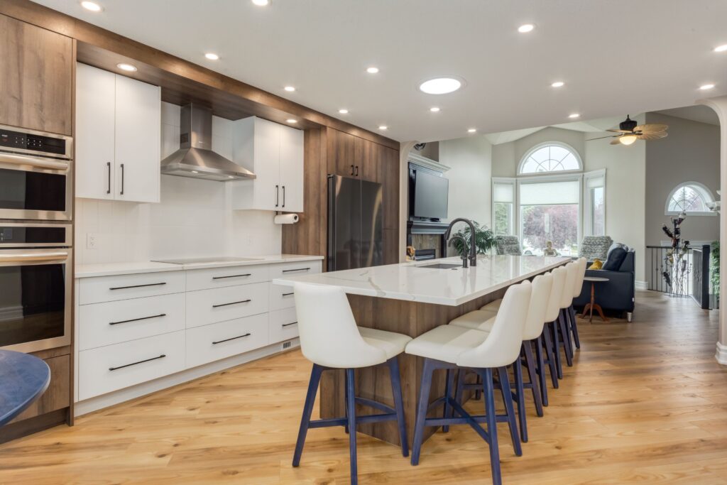 Modern kitchen with white cabinets, wooden accents, a large island with six white chairs, stainless steel appliances, wood flooring, and an open layout leading to a bright living area with large windows.