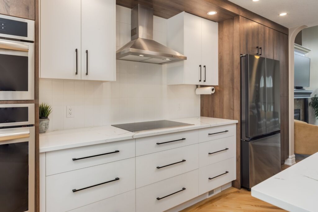 Modern kitchen with white cabinets, black handles, an electric cooktop, stainless steel range hood, double wall ovens, a black refrigerator, wood accents, and under-cabinet lighting.