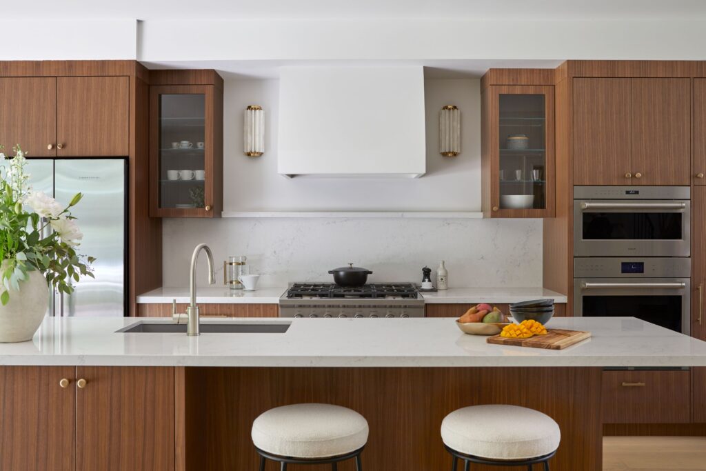 Modern kitchen with wood cabinets, white countertops, stainless steel appliances, and a central island with two round cushioned stools. A vase of flowers and a fruit bowl decorate the island.