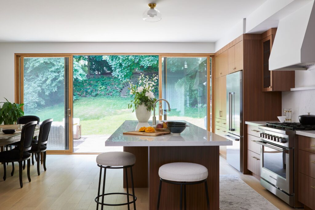 Modern kitchen with wood cabinets and a large island with two stools, facing floor-to-ceiling glass doors that overlook a green backyard. A dining table with black chairs sits to the left, and flowers decorate the island.