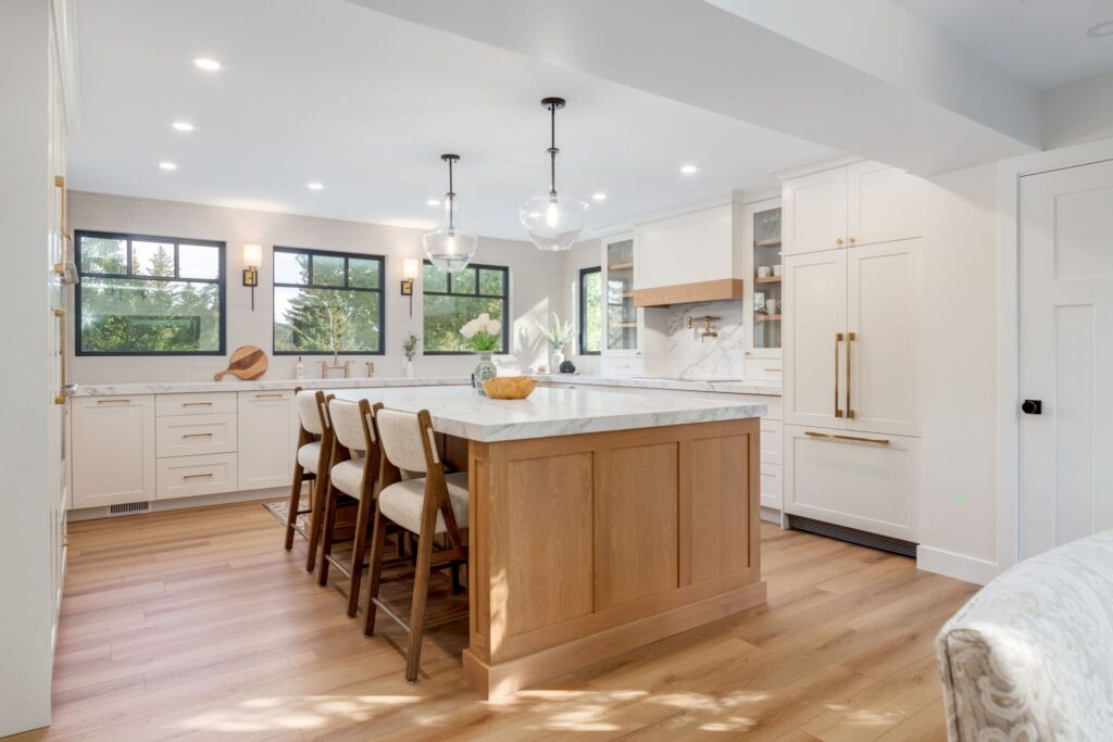 Bright, modern kitchen with white cabinets, marble countertops, a large island with wooden barstools, pendant lights, light wood floors, and large windows letting in natural light.