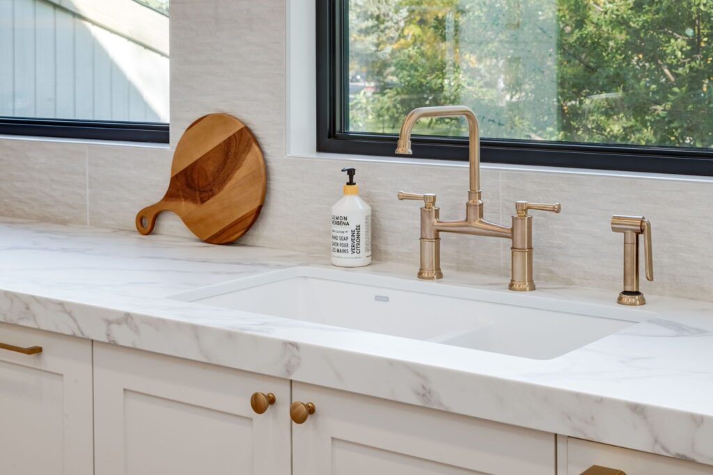 Modern kitchen sink with a white marble countertop, brushed gold faucet, soap dispenser, and a wooden cutting board resting against a tiled backsplash beneath a large window with a view of trees.