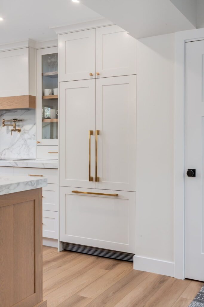 A modern kitchen with light wood flooring, white cabinets, gold handles, marble backsplash, and a built-in refrigerator with matching cabinet panels and handles.