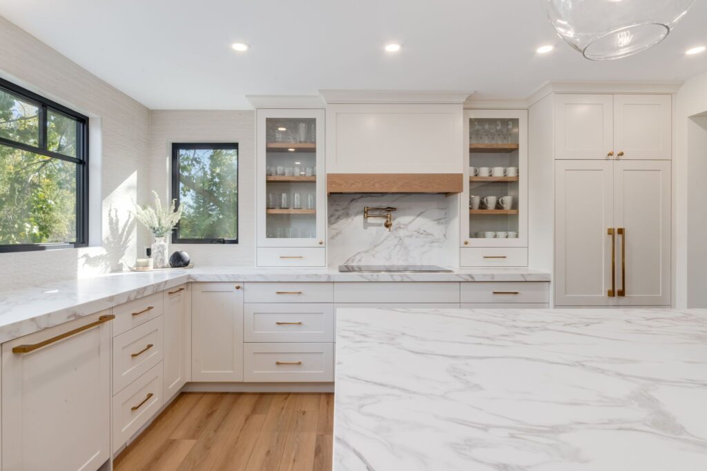 Bright, modern kitchen with white cabinets, marble countertops, gold handles, wood floors, and large windows letting in natural light. Glass-front cabinets display dishes and a wooden range hood is above the stove area.