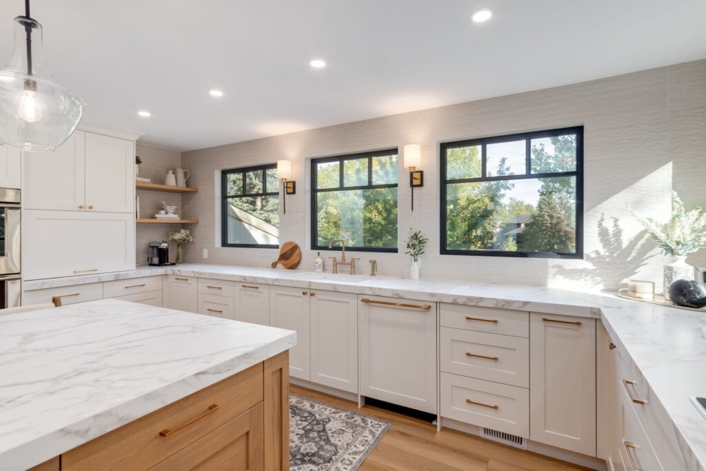 Bright, modern kitchen with white cabinets, marble countertops, large island with wooden base, two windows letting in natural light, and decorative items on the counters and shelves.