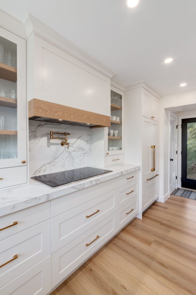 Modern kitchen with white cabinets, marble countertops and backsplash, brass hardware, built-in stove, glass-front cabinets, wooden floor, and a black-framed glass door leading outside.