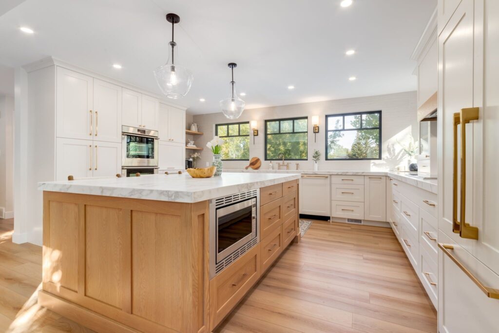 Modern kitchen with light wood floors, a large wood and white marble island, built-in oven, microwave, white cabinets with gold handles, pendant lights, and large windows letting in natural light.