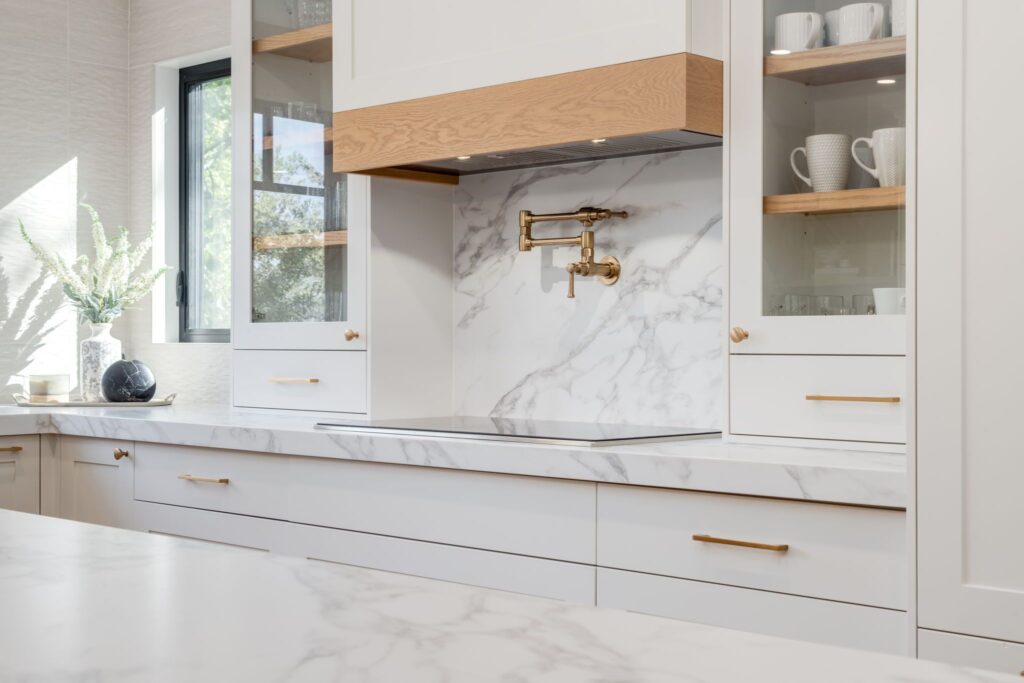 Modern kitchen with white cabinets, gold hardware, marble countertops and backsplash, a gold pot filler, glass-front cabinets with dishes, a window, and a vase with greenery on the counter.