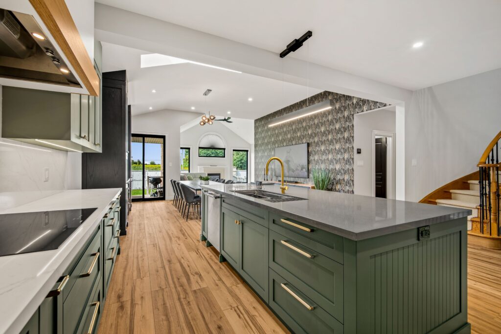 Modern open-concept kitchen with green island, gold faucet, and gray countertops, leading into a dining and living area with wood flooring, patterned accent wall, and large windows letting in natural light.