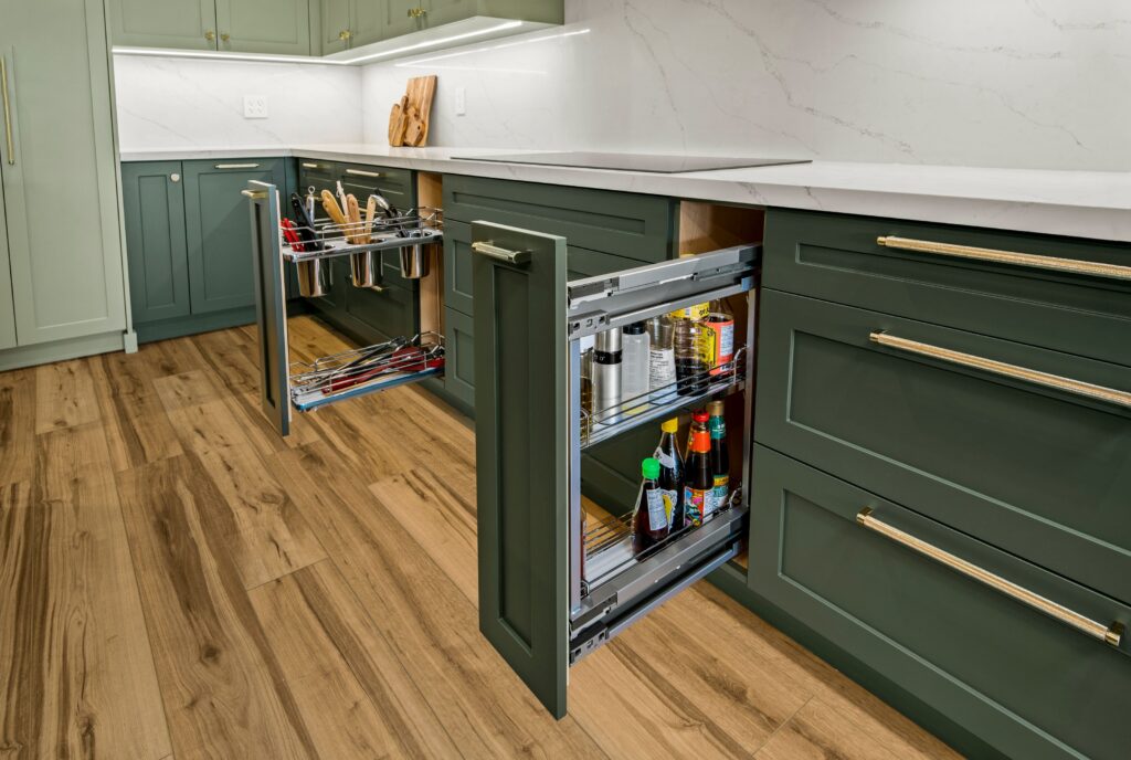 Modern kitchen with green cabinets and wood flooring, featuring two pull-out pantry drawers open to reveal organized cooking utensils, canned goods, and bottles inside. White countertops and marble backsplash are visible.