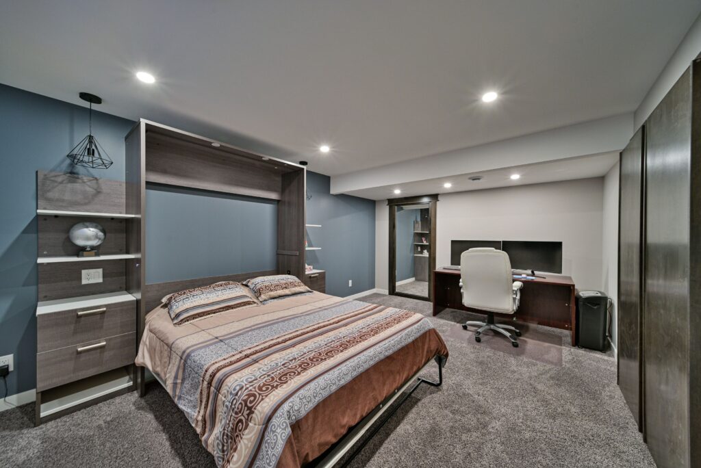 A modern bedroom with a double bed, patterned bedding, bedside shelves, and a desk with a computer and white chair in a carpeted room with blue and gray walls and recessed lighting.