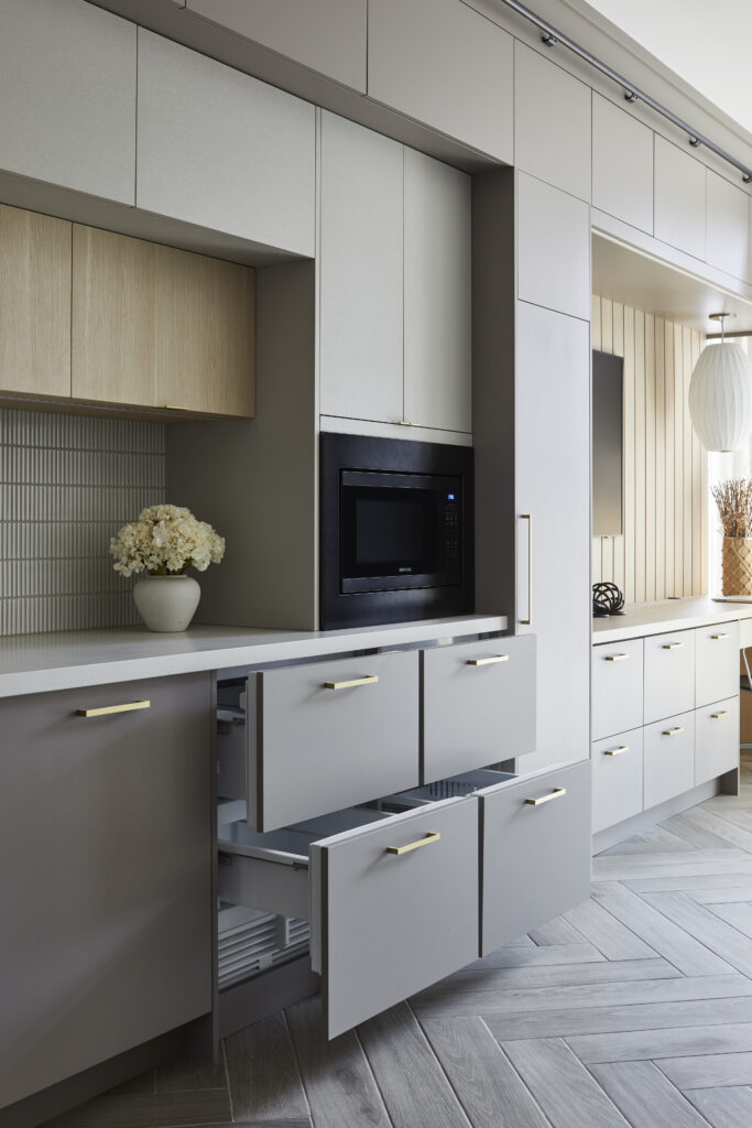 Modern Toronto condo kitchen renovation featuring gray cabinets with gold handles, two open drawers, built-in microwave, light wood accents, a vase of white flowers on the counter, and stylish decorative objects in the background.