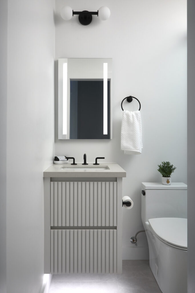 A modern bathroom with a white vanity, rectangular mirror with vertical lights, black fixtures, a towel ring with a white towel, a toilet, and a small potted plant on the toilet tank—perfect inspiration for your Toronto condo kitchen renovation.
