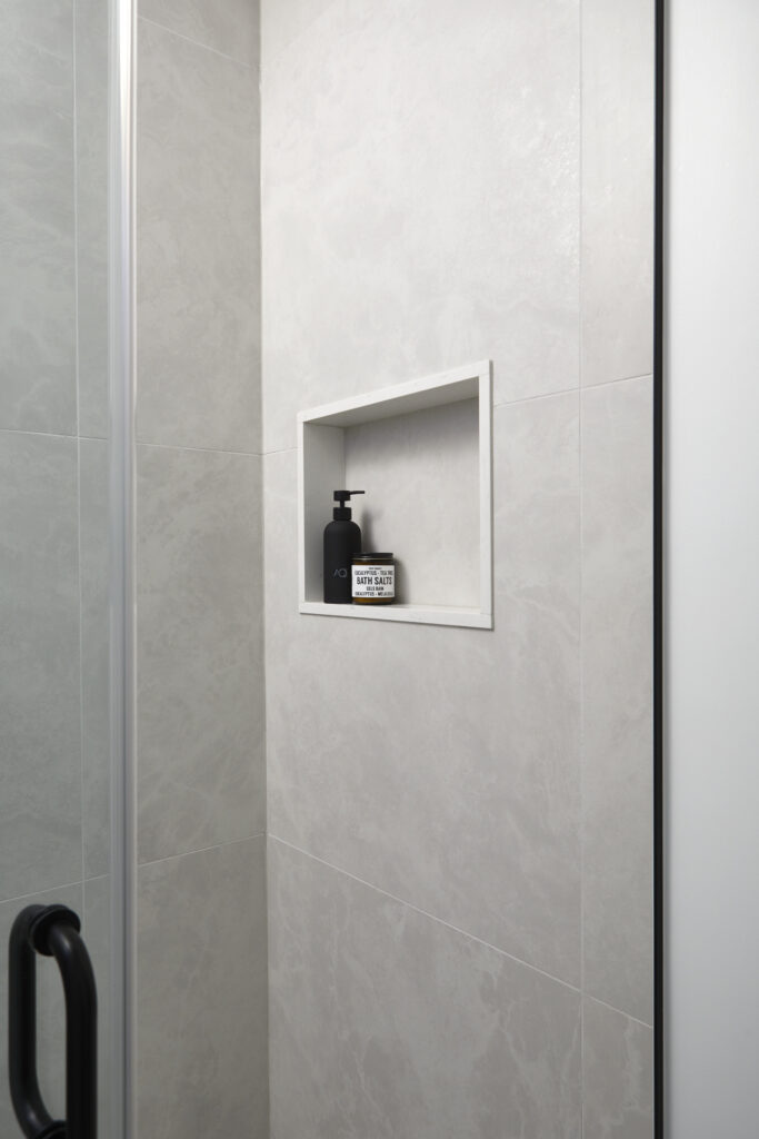 A built-in shower niche with a black soap dispenser and a small jar on a white shelf, set against light gray tiled walls, mirrors the sleek style often seen in Toronto condo kitchen renovation projects. A black shower door handle is visible on the left side.