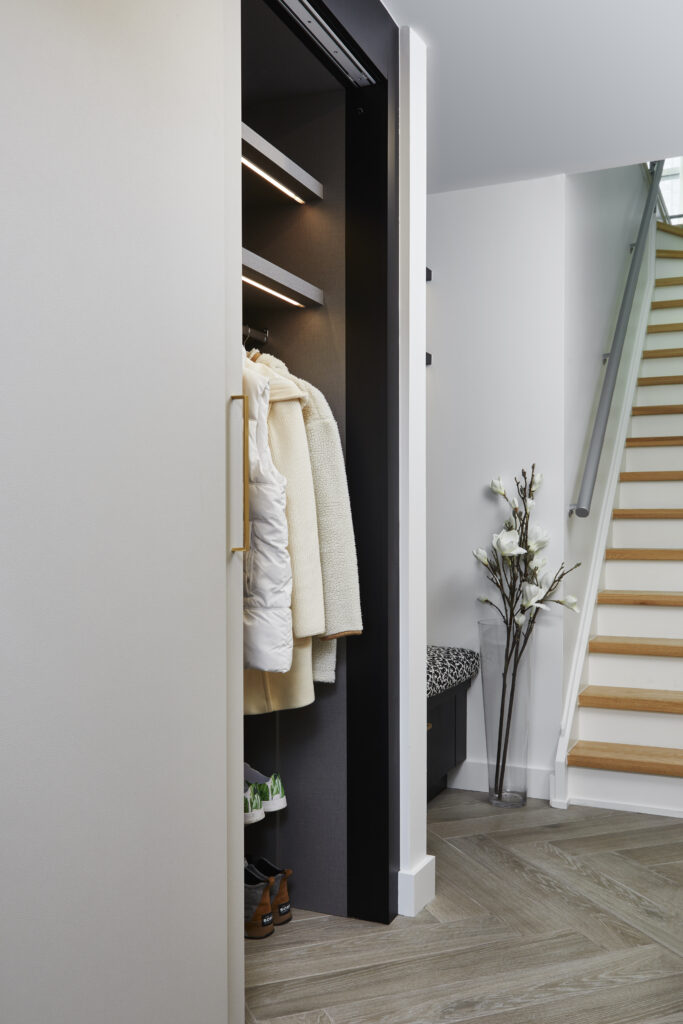 A modern hallway with light wood floors, an open closet holding white and beige coats, a bench with a patterned cushion, floral arrangement, and a staircase with wooden steps and metal railing—perfectly complementing a Toronto condo kitchen renovation.
