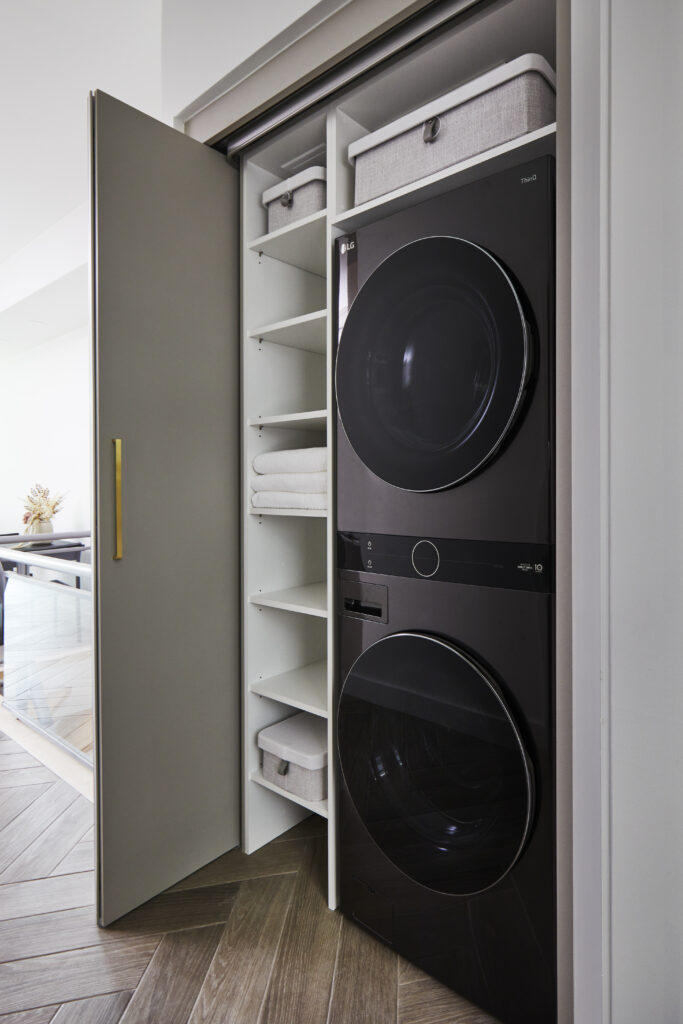 A modern laundry closet, inspired by Toronto condo kitchen renovation style, featuring a stacked black washer and dryer, open shelves with baskets and folded towels, and sliding doors with a gold handle.