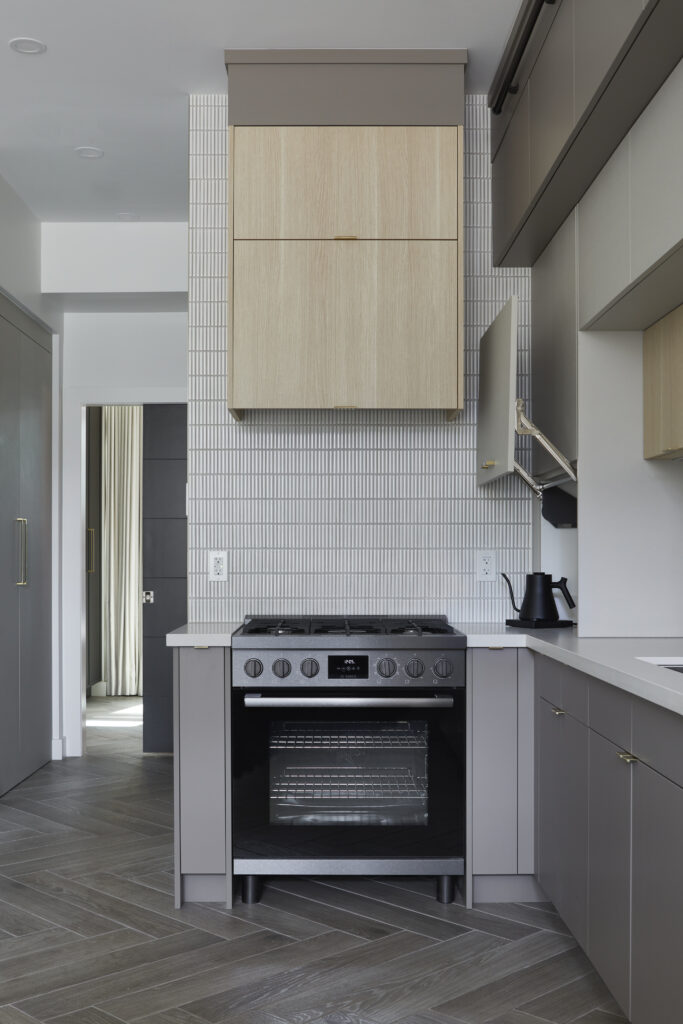 Modern Toronto condo kitchen renovation featuring a built-in oven and range, light wood and gray cabinets, white tiled backsplash, herringbone-patterned gray floor, and a black electric kettle on the counter.