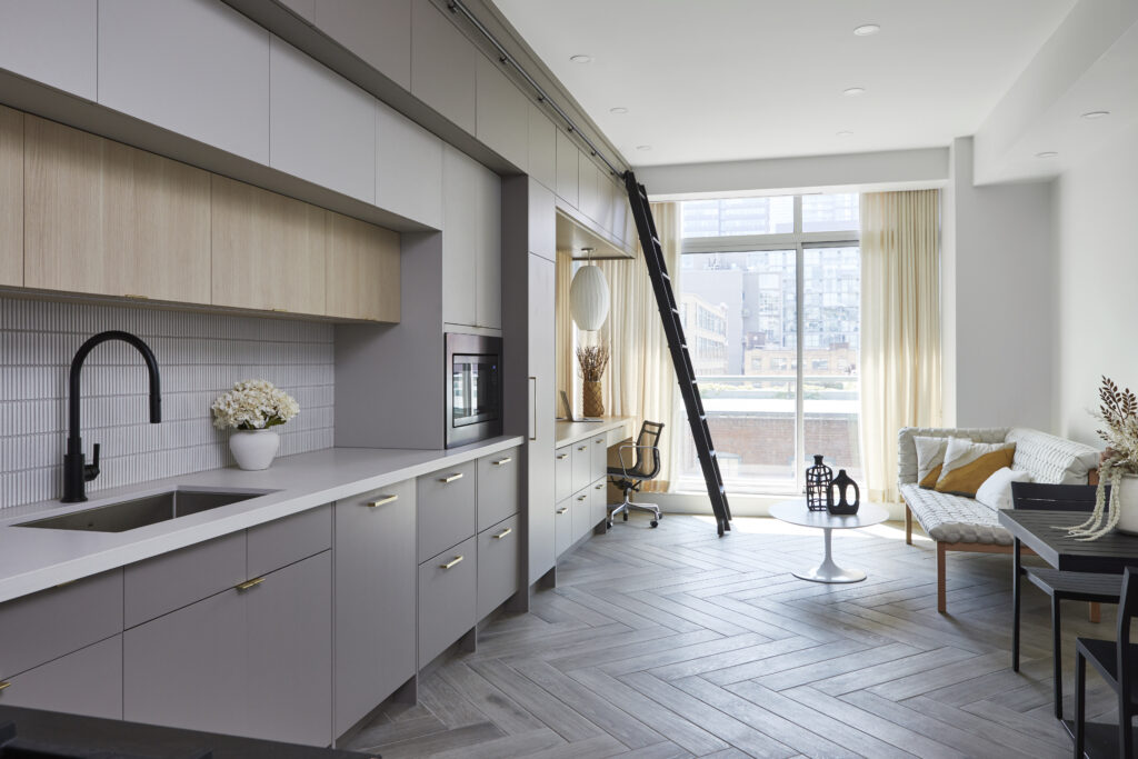 Modern apartment featuring a sleek gray Toronto condo kitchen renovation, built-in cabinets, black faucet, and herringbone wood floors. Enjoy a cozy sitting area, large windows with city views, and a tall ladder on a rail by the desk.