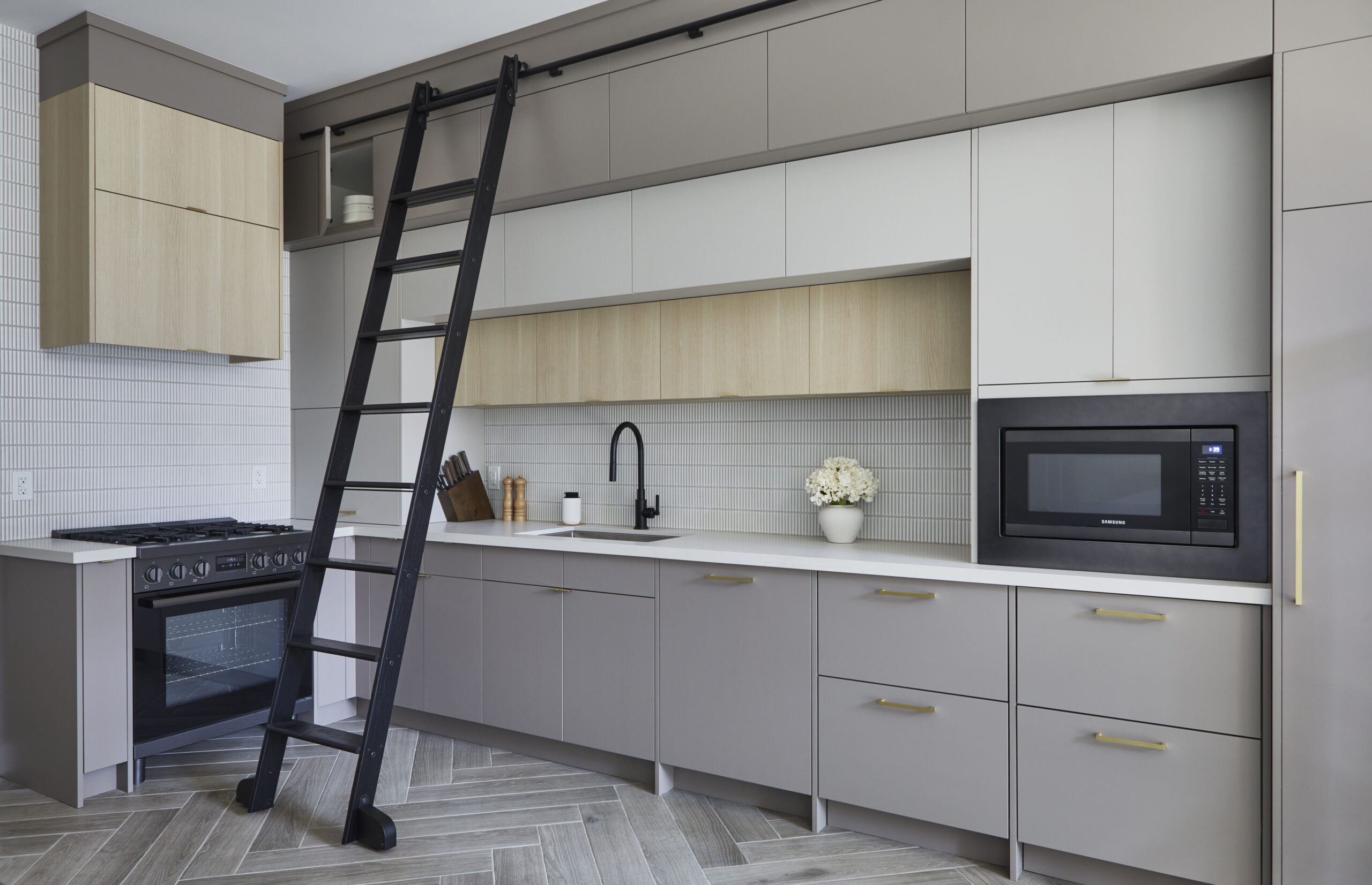 Modern Toronto condo kitchen renovation featuring gray and beige cabinets, a black stove, microwave, sleek black sink, and sliding ladder for upper cabinet access. Herringbone floor and white backsplash complete the look.