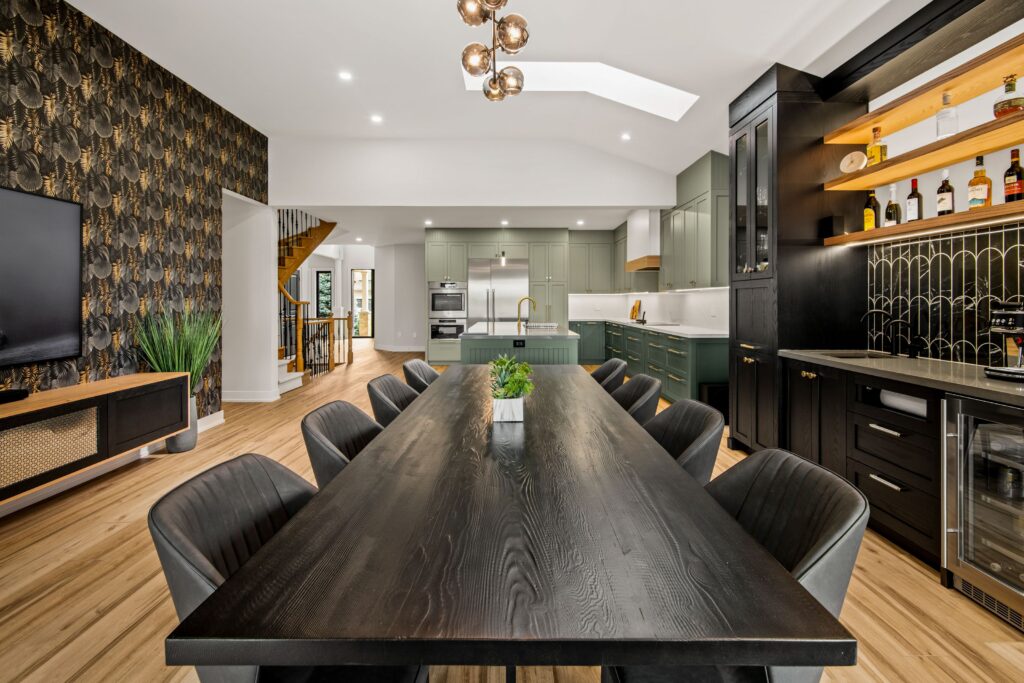 Modern dining room and kitchen with a long black dining table, black chairs, green cabinetry, wooden flooring, stylish lighting, and a black accent bar area with shelves of bottles. A patterned wall and plants add decorative touches.