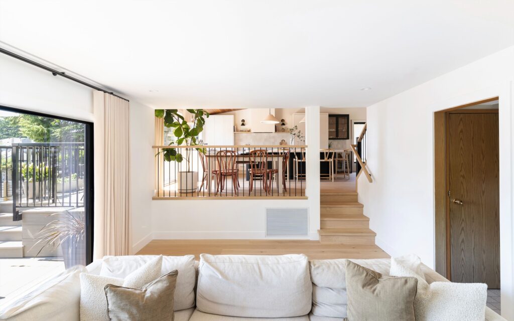 A modern living room with a beige sofa in the foreground, light wood floors, and stairs leading up to a dining area with wooden chairs and a large plant. Large windows let in natural light.