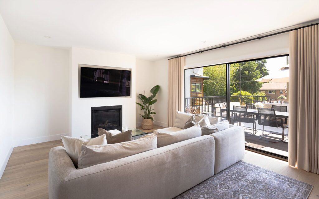 A modern living room with a beige sectional sofa, wall-mounted TV above a fireplace, glass coffee table, potted plant, large sliding doors, and a view of an outdoor patio and greenery.