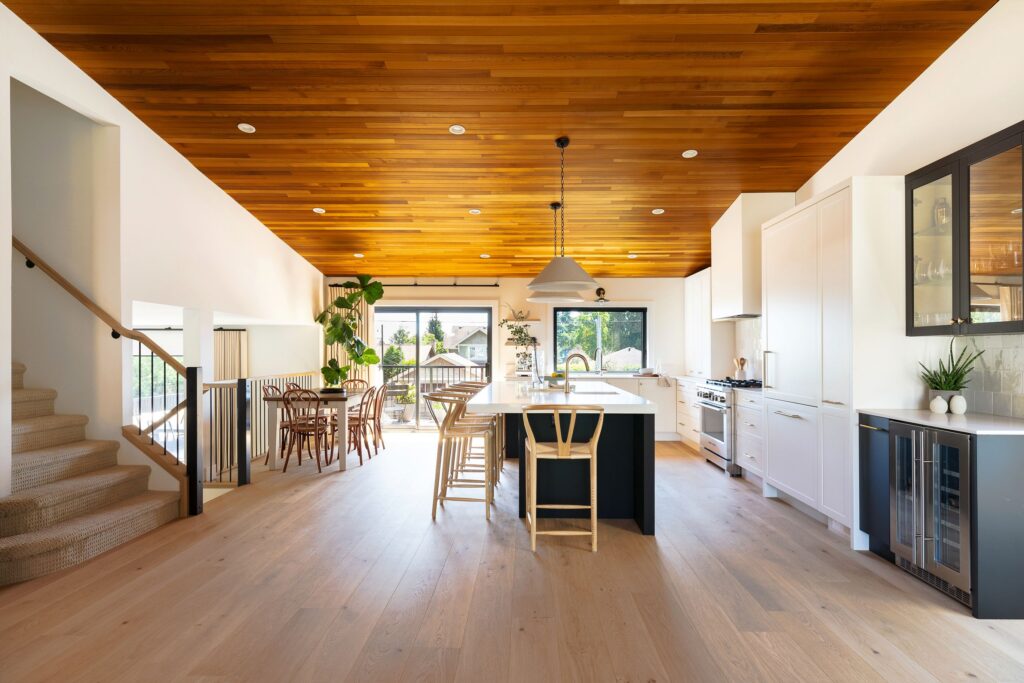 Modern kitchen and dining area with light wood floors and a wooden ceiling. White cabinets, a large island with barstools, pendant lighting, and large windows letting in natural light. A staircase is visible on the left.