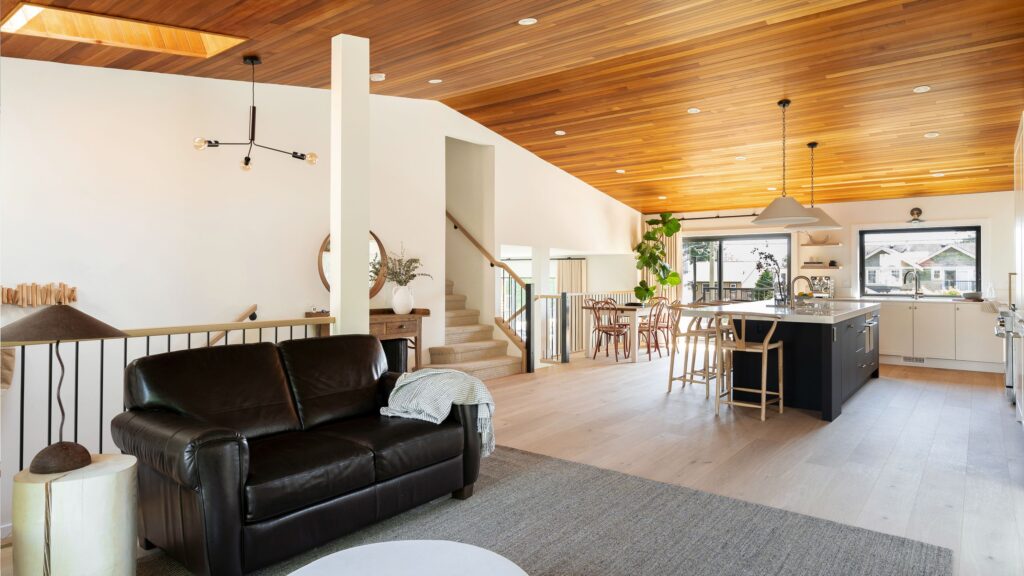 Modern open-concept living space with a black leather sofa, wood ceiling, light wood floors, a rug, staircase, and a kitchen area featuring a large island, bar stools, pendant lighting, and large windows.