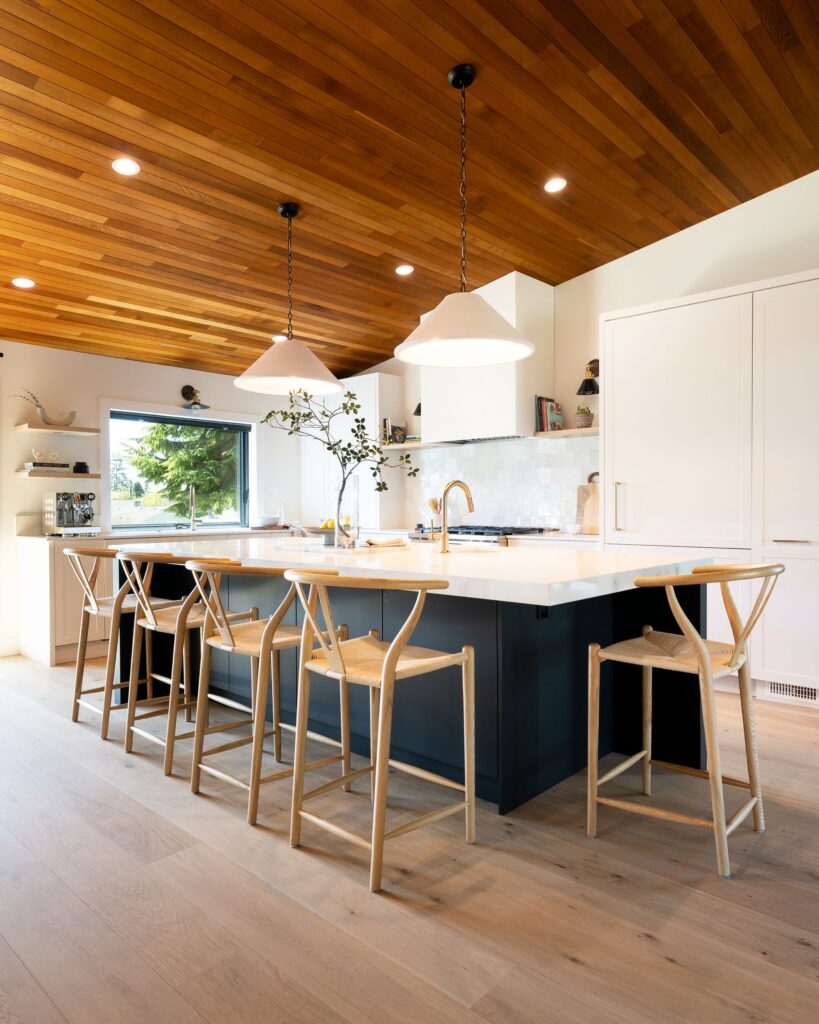 Modern kitchen with a wood-paneled ceiling, large island with a white countertop, four wooden bar stools, pendant lights, white cabinets, and a window letting in natural light.