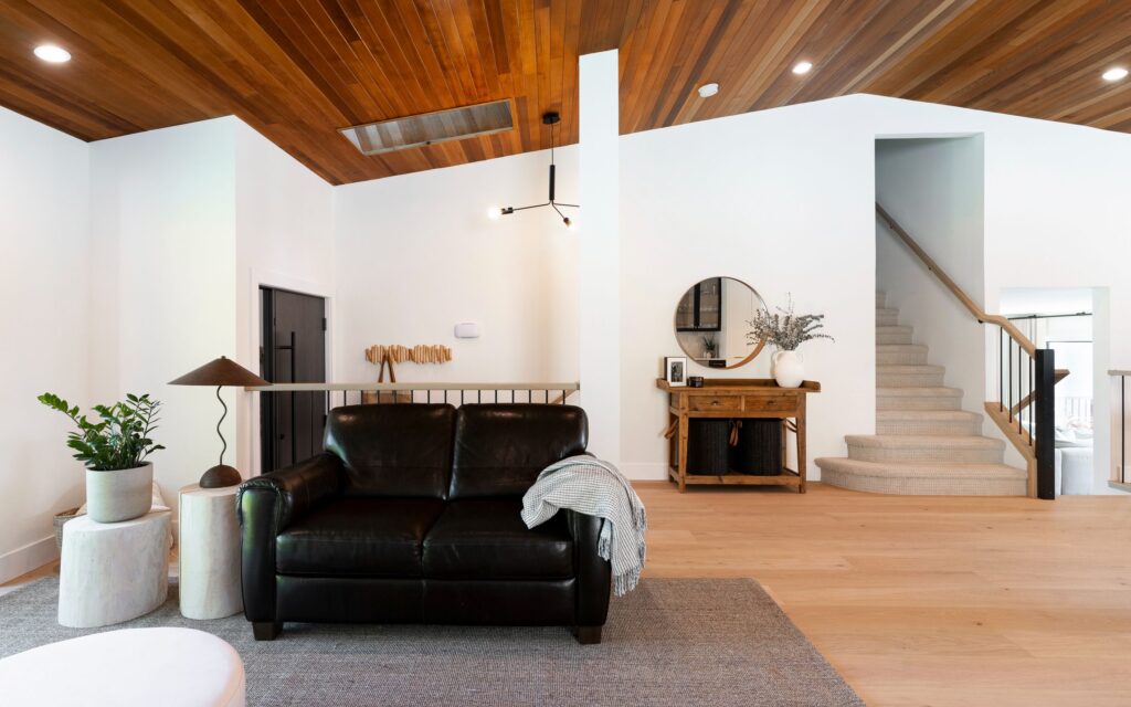 Modern living room with a black leather loveseat, two white side tables with potted plants, a wooden console with a round mirror, and carpeted stairs leading up. The ceiling is wooden and the floor is light wood.