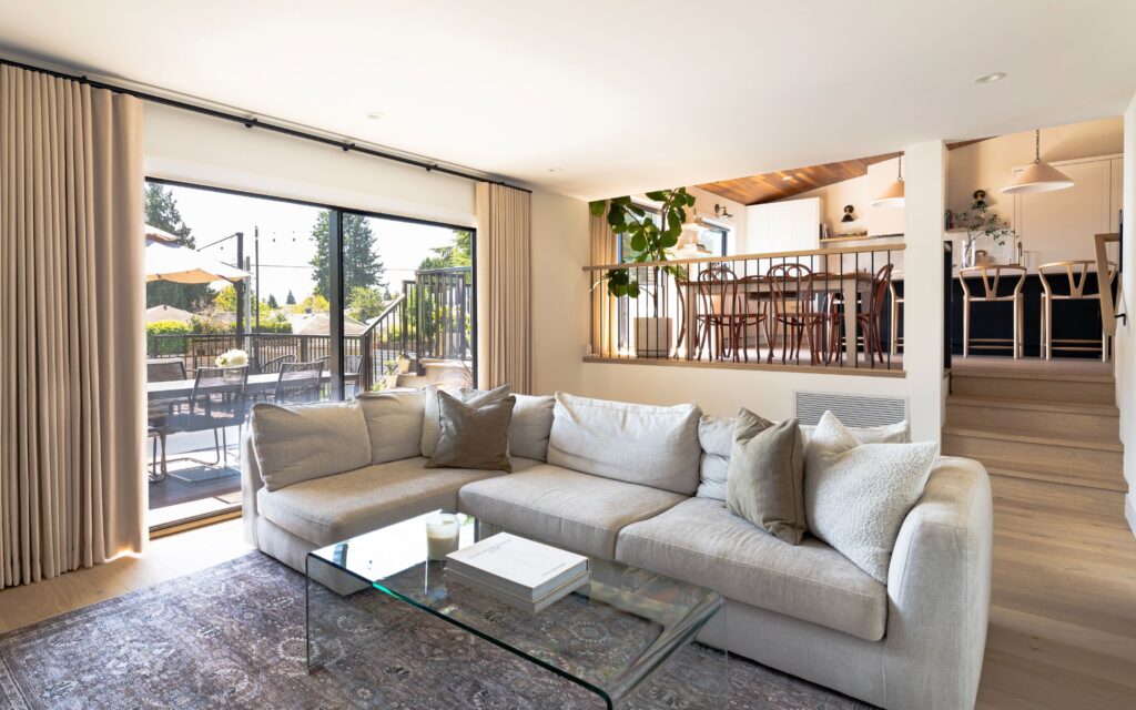 A bright, modern living room with a large beige sectional sofa, glass coffee table, and rug, opening to a patio through sliding glass doors. An elevated dining area with wooden chairs is visible in the background.