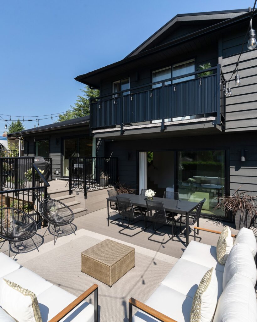 A modern outdoor patio with cushioned seating, a wicker table, black dining set, string lights, and dark gray house exterior under a clear blue sky.