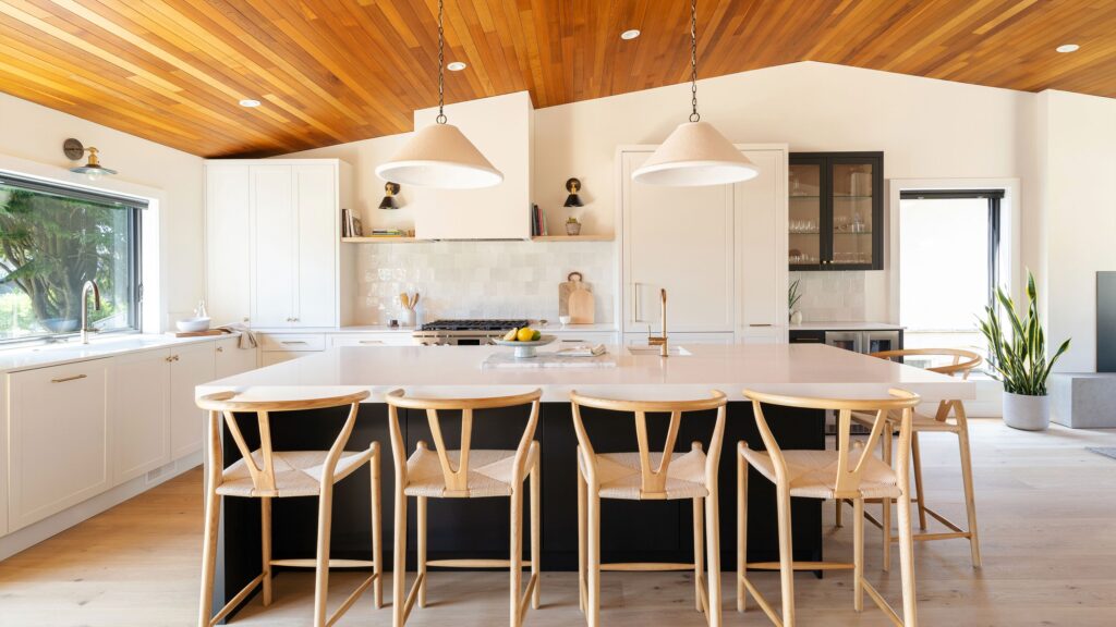 Modern kitchen with a large island, four wooden chairs, white cabinetry, pendant lights, a wood-paneled ceiling, and a large window letting in natural light. There’s a plant near the door and minimal, stylish decor.