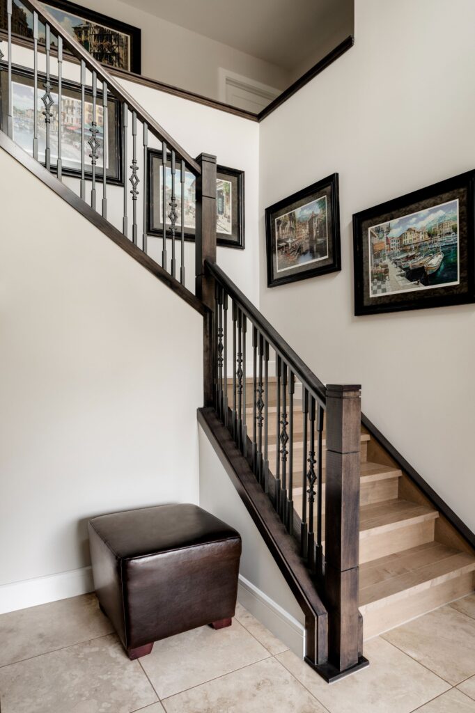 A wooden staircase with black metal railings leads up next to a white wall decorated with framed paintings. A dark brown leather ottoman sits at the base on beige tiled flooring.