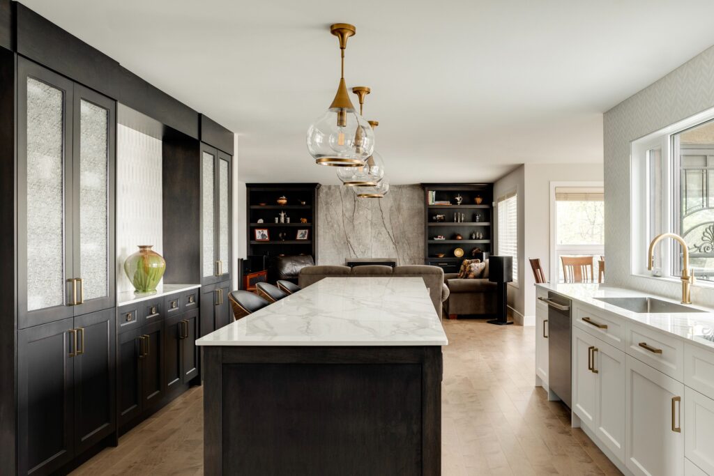 Modern kitchen with a marble island, black cabinets, gold pendant lights, and white countertops. An open living area with a stone accent wall and shelves is visible in the background. Large window lets in natural light.