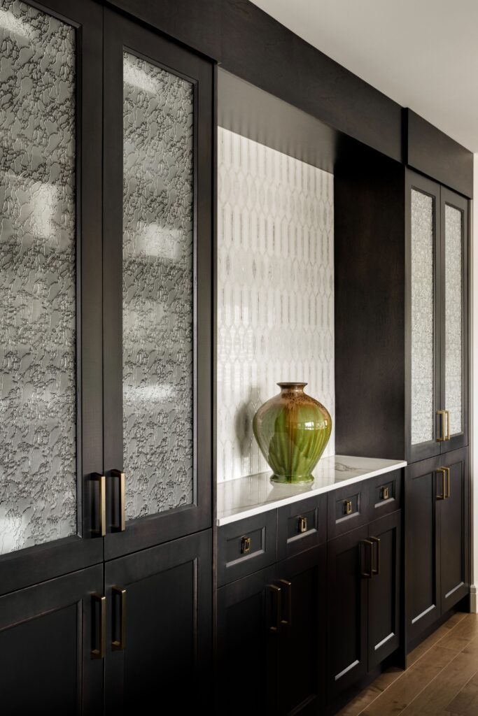 Dark wood cabinets with frosted glass panels flank a white patterned backsplash. A large green and brown ceramic vase sits on a white countertop, and brass hardware accents the cabinetry.