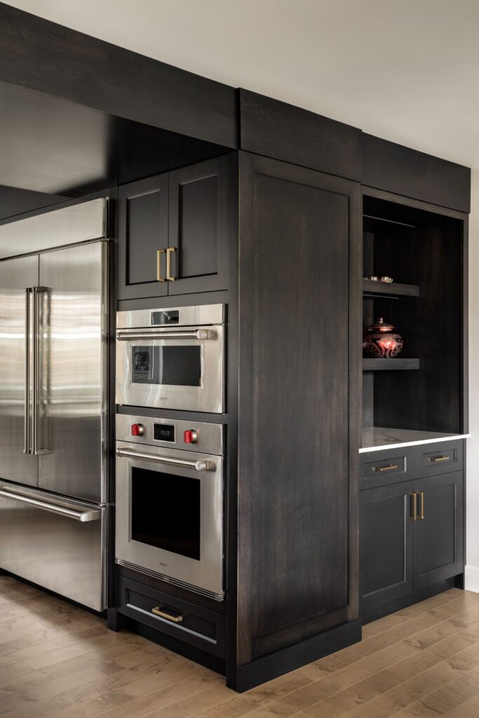 Modern kitchen with dark wood cabinets, built-in stainless steel double ovens, and a large stainless steel refrigerator. Open shelving displays decorative items; lower cabinets have gold handles. Light wood floor contrasts with dark cabinetry.