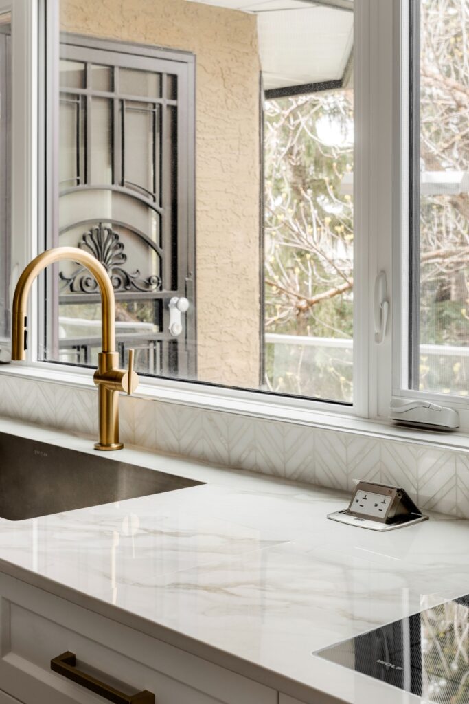 A modern kitchen countertop with a gold faucet, under-mount sink, marble surface, geometric tile backsplash, and an electrical outlet. A large window overlooks an outdoor scene with trees and a metal door.