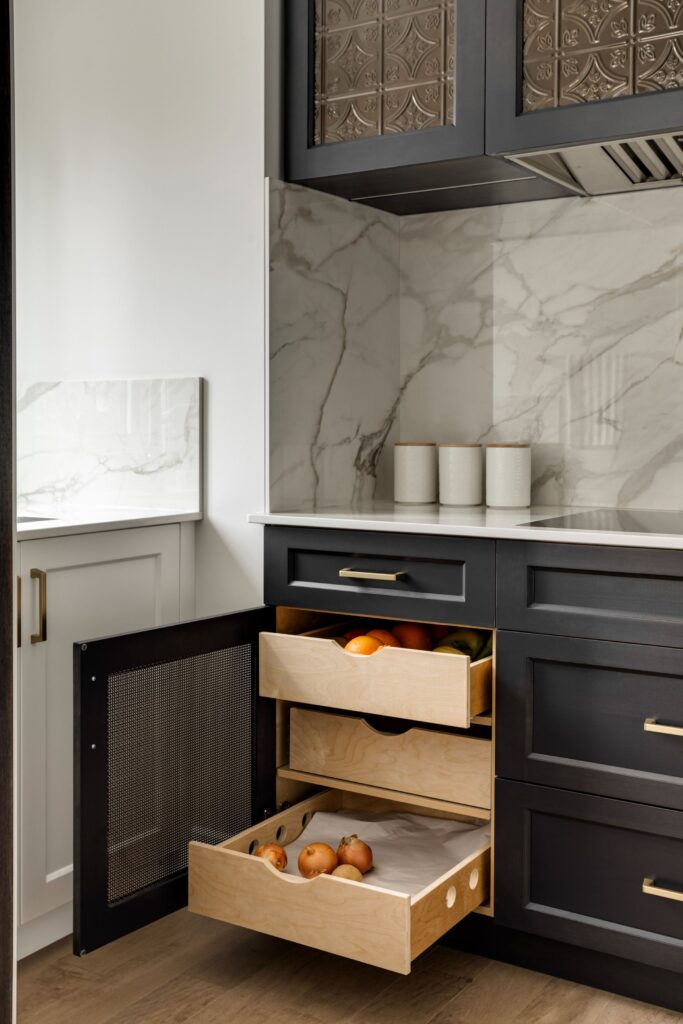 Modern kitchen with black cabinets, marble backsplash, and two open wooden drawers holding fresh produce like onions and apples. Three white canisters sit on the countertop. The floor is wood.