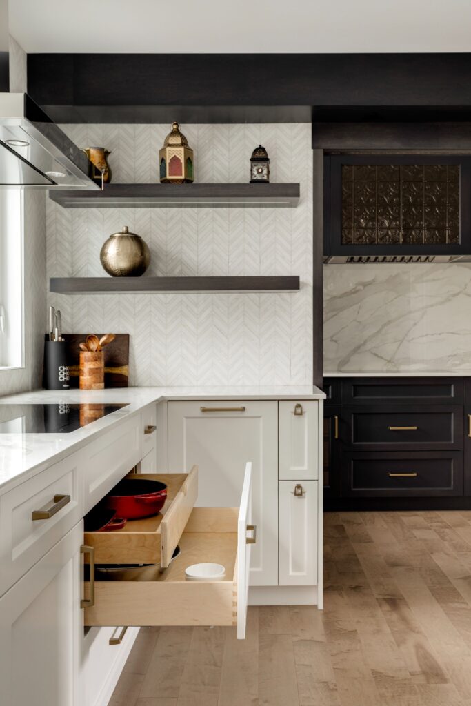 Modern kitchen with white cabinets, light wood flooring, open shelves with decorative items, and a drawer slightly open showing red cookware. The backsplash features a subtle herringbone pattern. Black cabinetry frames the stove area.